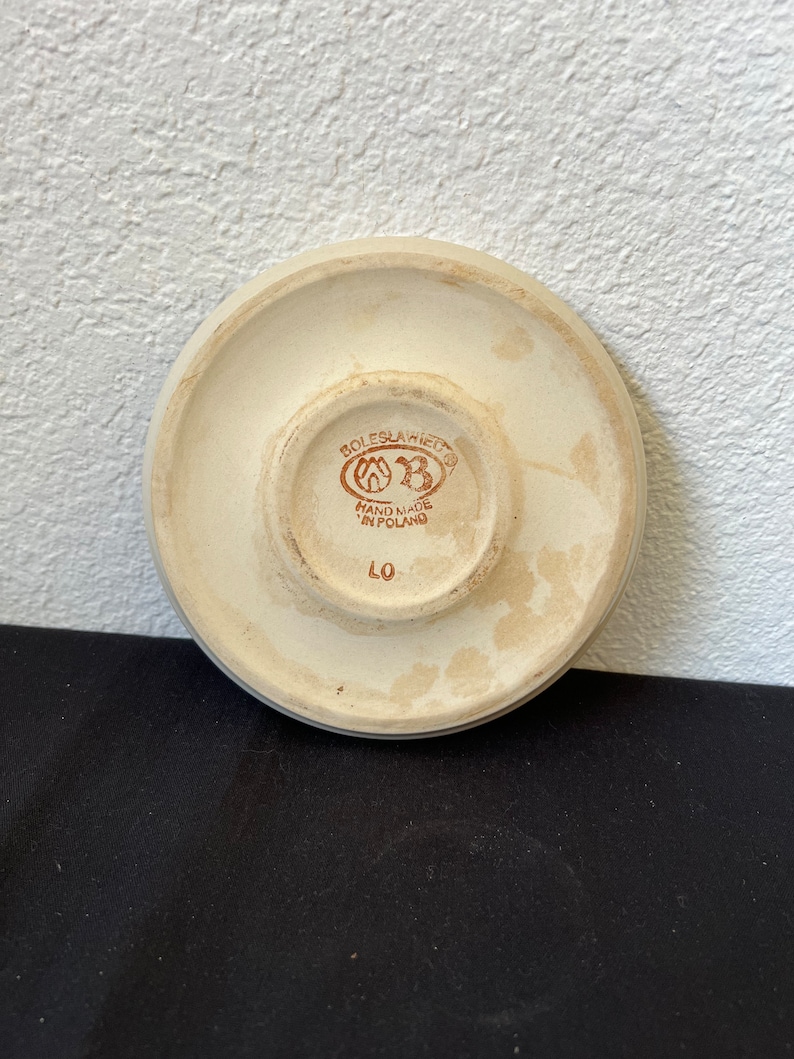 Puede incluir: Parte inferior de un platillo de cer&aacute;mica color crema con un borde elevado. El centro presenta un emblema marr&oacute;n con el texto "BOLESLAWIEC" y "HAND MADE IN POLAND". Tambi&eacute;n es visible el n&uacute;mero "LO".