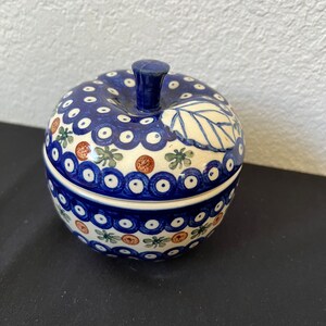 Puede incluir: Una caja de baratijas de cer&aacute;mica con forma de manzana con un patr&oacute;n floral azul y blanco. La tapa tiene un tallo azul oscuro y un dise&ntilde;o de hoja. La base de la caja tiene un patr&oacute;n similar con acentos marrones y verdes. La caja est&aacute; sobre una superficie negra.