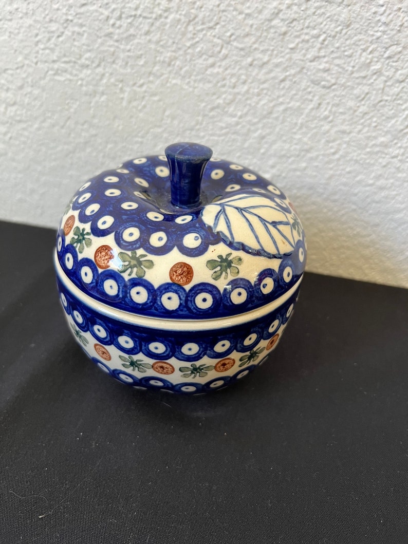 Puede incluir: Una caja de baratijas de cer&aacute;mica con forma de manzana con un patr&oacute;n floral azul y blanco. La tapa tiene un tallo azul oscuro y un dise&ntilde;o de hoja. La base de la caja tiene un patr&oacute;n similar con acentos marrones y verdes. La caja est&aacute; sobre una superficie negra.