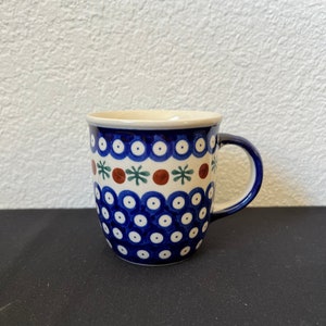 Puede incluir: Una taza de cer&aacute;mica con un patr&oacute;n de lunares azules y blancos, acentuada con dise&ntilde;os florales marrones y verdes. La taza tiene un asa azul y un borde color crema. La taza probablemente se usa para beber bebidas.