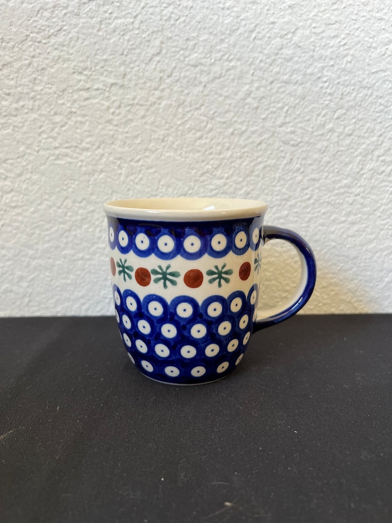 Puede incluir: Una taza de cer&aacute;mica con un patr&oacute;n de lunares azules y blancos, acentuada con dise&ntilde;os florales marrones y verdes. La taza tiene un asa azul y un borde color crema. La taza probablemente se usa para beber bebidas.