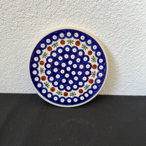 Puede incluir: Un posavasos redondo de cer&aacute;mica con un estampado floral azul y blanco. El posavasos presenta un borde blanco y un dise&ntilde;o de c&iacute;rculos azules, puntos marrones y formas de estrella verdes. El posavasos est&aacute; sobre una superficie negra.