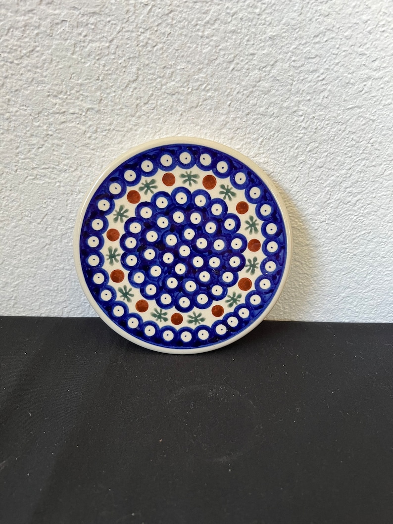 Puede incluir: Un posavasos redondo de cer&aacute;mica con un estampado floral azul y blanco. El posavasos presenta un borde blanco y un dise&ntilde;o de c&iacute;rculos azules, puntos marrones y formas de estrella verdes. El posavasos est&aacute; sobre una superficie negra.