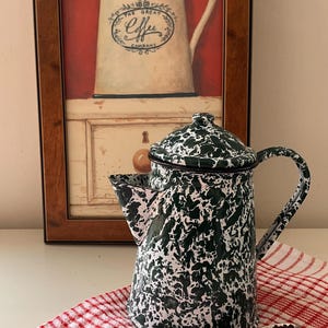 Peut inclure: Une cafetière en émail moucheté vert et blanc avec un couvercle et une anse assortis. Une cuillère en argent remplie de marc de café brun foncé repose sur un tissu à carreaux rouges et blancs devant la cafetière, avec une affiche encadrée.