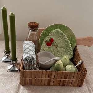 May include: A wicker basket filled with green and white ceramic dishes, a clear glass bottle with a cork stopper, and green ceramic salt and pepper shakers. The basket is sitting on a white tablecloth.