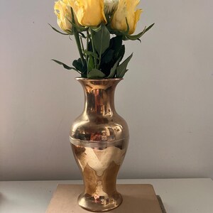 Peut inclure: Un vase en laiton avec une base arrondie et un col étroit, contenant un bouquet de roses jaunes. Le vase est posé sur deux livres, l'un intitulé "C.W. Anderson's Favorite Horse Stories" et l'autre intitulé "Textile Fibers and Their Use".
