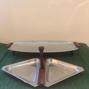 Peut inclure: Deux plateaux de service vintage. L'un est de forme ovale allongée avec des poignées et des pieds en bois. L'autre est de forme triangulaire avec deux compartiments et une poignée en bois. Les deux plateaux sont de couleur argent.