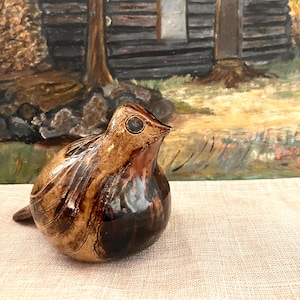 May include: A ceramic bird ornament with a glossy finish. The bird is mainly brown with darker brown detailing and a dark eye. The ornament is set against a backdrop of a painting depicting a rustic cabin in a forest setting.