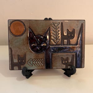 May include: A rectangular ceramic tile with a cat theme. The tile features a large cat face, several smaller cat figures, and a sun. The colour palette includes brown, blue, and orange.