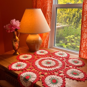 May include: A decorative, red and white crochet doily with a floral pattern is displayed on a wooden table. The doily features a central circular design and eight flower-shaped motifs with green centers. A lamp and vase with pink flowers are also on the table.