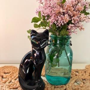May include: A glossy black ceramic cat figurine with stylised features sits beside a translucent teal glass mason jar filled with a bouquet of light purple lilac flowers. The cat has a smooth, reflective surface and a pointed ear silhouette.