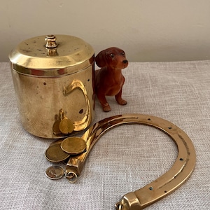 Peut inclure: Ensemble d'objets en laiton vintage : un pot avec couvercle, une poignée en forme de fer à cheval et une figurine de teckel. Plusieurs pièces de monnaie sont placées près de la poignée. Les objets sont disposés sur une surface de ton neutre.