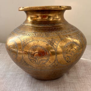 Peut inclure: Un pot en laiton vintage avec des motifs floraux et géométriques gravés. Le pot a un corps arrondi, un bord évasé et un col légèrement resserré. Le laiton a une teinte dorée chaude avec des zones de patine.