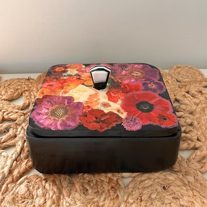 Peut inclure: Une boîte carrée noire avec un couvercle orné d'un motif floral. Le couvercle présente un collage de fleurs rouges, oranges et violettes. Un bouton géométrique blanc et noir sert de poignée. La boîte est probablement pour le rangement.