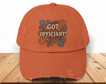 May include: An orange distressed baseball cap with the text "GOT OFFICIANT?" in a retro floral design. The design includes blue, orange, and yellow flowers. The cap has a curved brim and a distressed look.