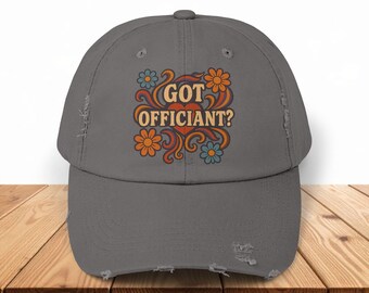 May include: Distressed gray baseball cap with the text "GOT OFFICIANT?" in a retro floral design. The design features orange, blue, and yellow flowers with a swirl pattern. The cap has a pre-worn look.