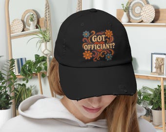 May include: Black distressed baseball cap with the text "GOT OFFICIANT?" in orange and blue retro-style lettering, surrounded by floral designs. The cap has a curved brim and a worn appearance.