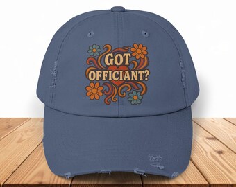 May include: A distressed blue baseball cap with the text "GOT OFFICIANT?" in a retro font, surrounded by orange and blue flowers. The cap has a pre-worn look with distressed edges, perfect for casual wear.