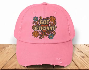 May include: A distressed pink baseball cap with the text "GOT OFFICIANT?" in a retro floral design. The design includes orange, blue, and brown flowers. The cap is sitting on a wooden surface.