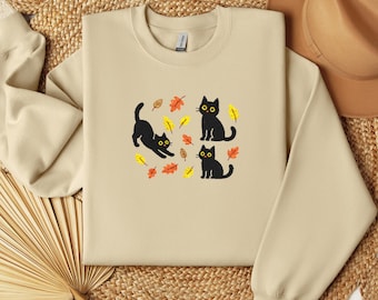 Puede incluir: Sudadera beige con un diseño de tres gatos negros con ojos amarillos y hojas de otoño. Los gatos están en diferentes poses, uno estirándose, otro sentado y otro de pie. La sudadera tiene cuello redondo y mangas largas.