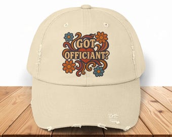 May include: A beige baseball cap with a distressed brim. The cap features a colorful floral design with the text "GOT OFFICIANT?" in orange and brown. The flowers are blue and orange.