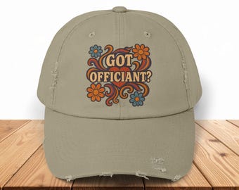May include: A tan baseball cap with a distressed brim. The cap features the text "GOT OFFICIANT?" in orange and white, surrounded by orange and blue flowers in a retro design. The hat is sitting on a wooden surface.