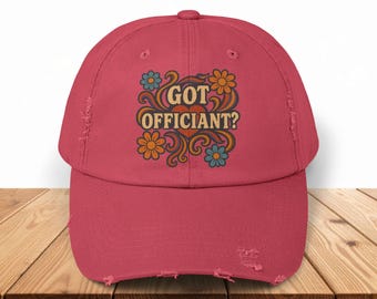 May include: A distressed red baseball cap with the text "GOT OFFICIANT?" in a retro floral design. The design features orange, blue, and yellow flowers. The cap has a curved brim and a worn appearance.