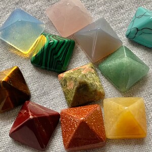May include: A set of ten pyramid-shaped crystals in various colors, including green, brown, yellow, red, and white. The crystals are arranged in a circle on a white fabric background.