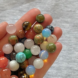 May include: A collection of small, round, polished stones in various colors, including green, blue, pink, brown, and white. The stones are arranged in a hand, with some overlapping.