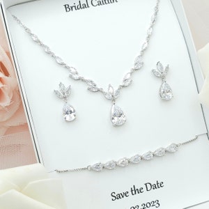 May include: A bridal jewelry set with a necklace, earrings, and bracelet. The set features a teardrop-shaped pendant with a cluster of smaller stones. The jewelry is silver-toned and has a clear crystal design. The box has the text "Save the Date 02.2023" printed on it.