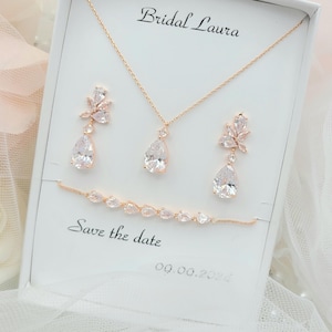 May include: A bridal jewellery set in rose gold featuring a necklace, earrings, and bracelet. The set is adorned with teardrop-shaped crystals and floral accents. The box displays the text "Bridal Laura" and "Save the date 09.00.2021".