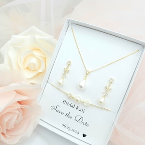 Puede incluir: Una caja blanca con un collar, pendientes y pulsera de oro con detalles de perlas. La caja tiene una tarjeta en el interior que dice "Bridal Kati Save the Date 06.25.2024".