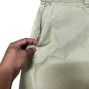Op de afbeelding: Een close-up van een lichtgroene broek met een gescheurde zak. De zak zit aan de rechterkant van de broek en wordt opengehouden door een hand.