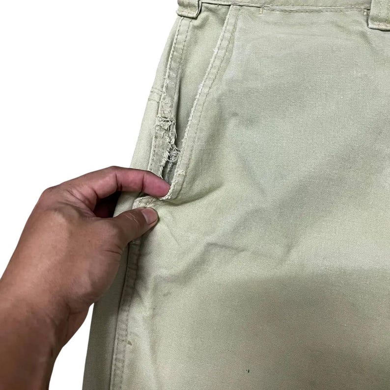 Op de afbeelding: Een close-up van een lichtgroene broek met een gescheurde zak. De zak zit aan de rechterkant van de broek en wordt opengehouden door een hand.