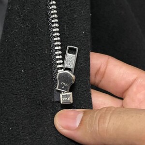 May include: Close-up of a black garment zipper with a silver metal pull. The zipper is partially closed, revealing the YKK brand name on the pull and the bottom stop. The fabric is a dark, textured material.