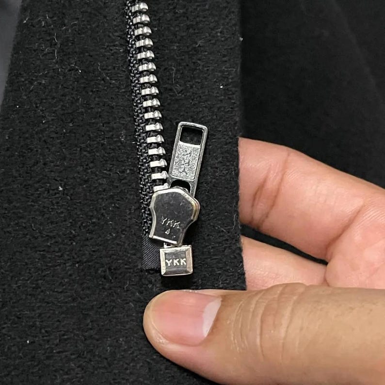 May include: Close-up of a black garment zipper with a silver metal pull. The zipper is partially closed, revealing the YKK brand name on the pull and the bottom stop. The fabric is a dark, textured material.