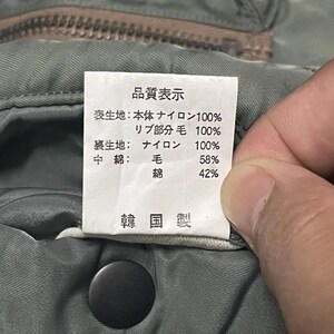 Puede incluir: Primer plano de una etiqueta con texto japon&eacute;s, sujeta por una mano, adjunta a una prenda verde. La etiqueta enumera la composici&oacute;n de la tela: 100% nailon, 58% lana y 42% algod&oacute;n. La prenda tiene una cremallera y un cierre a presi&oacute;n.