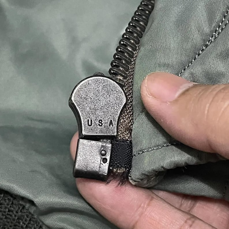 Puede incluir: Primer plano de un tirador de cremallera plateado con las letras "USA" estampadas. La cremallera est&aacute; sujeta a una tela verde, probablemente parte de una chaqueta o abrigo. Los dientes de la cremallera son negros.
