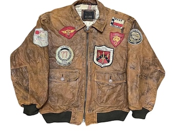 May include: A brown leather bomber jacket with multiple patches, including a Texas patch and a patch with the text "TASK FORCE 77". The jacket has a zip-up front, two front pockets, and a dark green ribbed collar and cuffs.