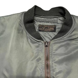 Puede incluir: Primer plano de una chaqueta bomber verde oliva con cuello y pu&ntilde;os de canal&eacute; negros. La chaqueta tiene cierre de cremallera completo y una etiqueta con texto. La etiqueta dice "JACKET FLYING MAN'S INTERMEDIATE".