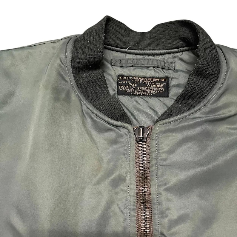 Puede incluir: Primer plano de una chaqueta bomber verde oliva con cuello y pu&ntilde;os de canal&eacute; negros. La chaqueta tiene cierre de cremallera completo y una etiqueta con texto. La etiqueta dice "JACKET FLYING MAN'S INTERMEDIATE".