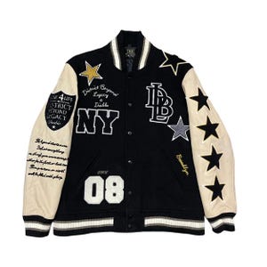 Op de afbeelding: Zwarte varsity jas met crèmekleurige mouwen en wit-zwarte gestreepte boorden. Voorzien van ster- en letterpatches, waaronder "NY" en "08". De jas heeft de tekst "District Beyond Legacy Diablo" en "Brooklyn".