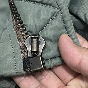 Puede incluir: Primer plano de una cremallera plateada en una chaqueta acolchada verde oliva. La cremallera est&aacute; parcialmente cerrada, revelando los dientes y el tirador met&aacute;lico. La tela tiene un patr&oacute;n de diamantes.