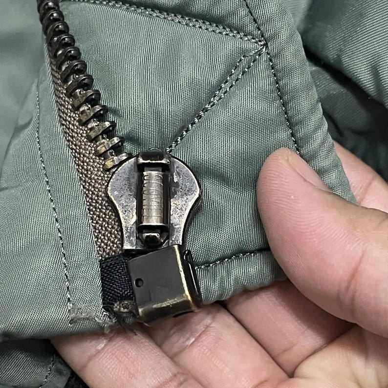 Puede incluir: Primer plano de una cremallera plateada en una chaqueta acolchada verde oliva. La cremallera est&aacute; parcialmente cerrada, revelando los dientes y el tirador met&aacute;lico. La tela tiene un patr&oacute;n de diamantes.