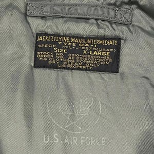 Puede incluir: Primer plano de una etiqueta de chaqueta verde militar. La etiqueta dice "JACKET FLYING, MAN'S, INTERMEDIATE" y "U.S. AIR FORCE". La etiqueta tambi&eacute;n incluye informaci&oacute;n sobre tallas, n&uacute;meros de stock y otros detalles.