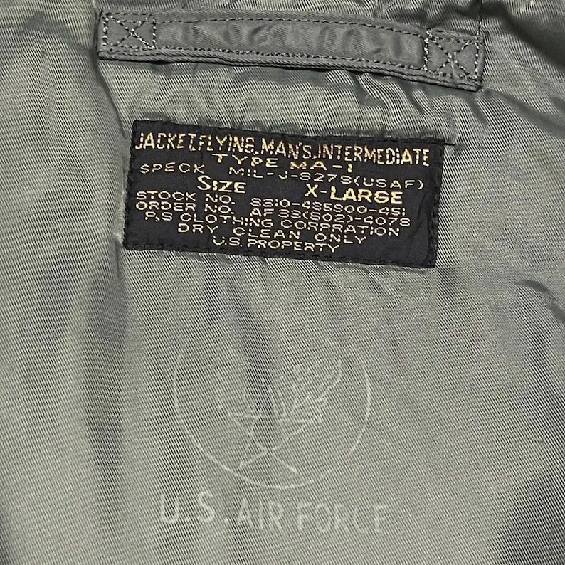 Puede incluir: Primer plano de una etiqueta de chaqueta verde militar. La etiqueta dice "JACKET FLYING, MAN'S, INTERMEDIATE" y "U.S. AIR FORCE". La etiqueta tambi&eacute;n incluye informaci&oacute;n sobre tallas, n&uacute;meros de stock y otros detalles.