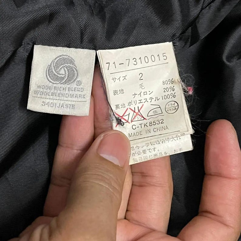 May include: Close-up of clothing tags with text. One tag reads "WOOL RICH BLEND WOOLBLENDMARK" and "$401JA978". The other tag has Japanese text, fabric percentages, and "MADE IN CHINA".