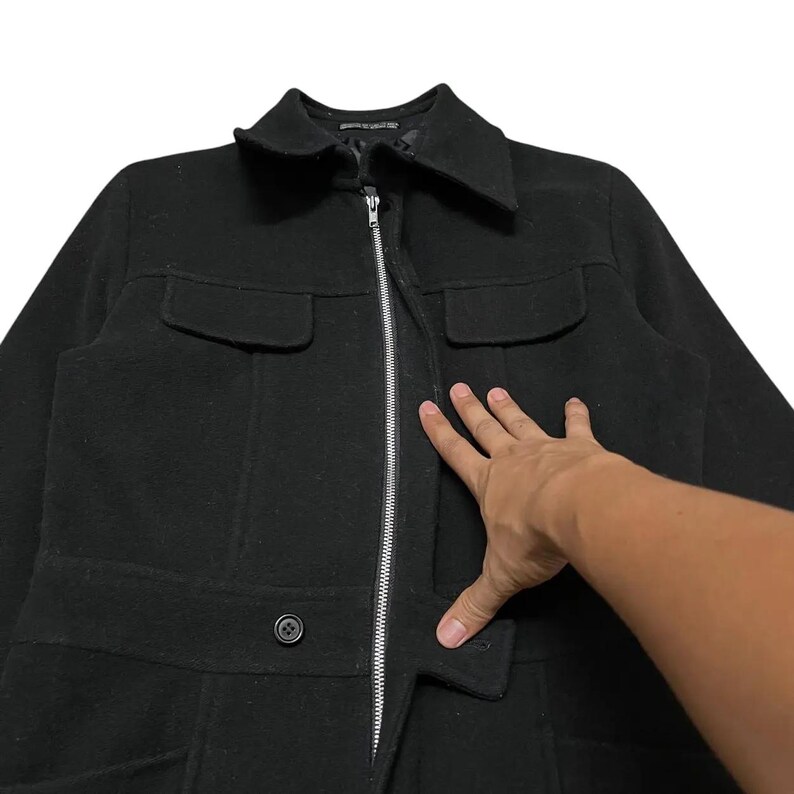 May include: Black wool coat with a collar, two chest pockets, and two lower pockets. The coat has a silver zipper closure and a button closure at the waist. The coat is a classic style.
