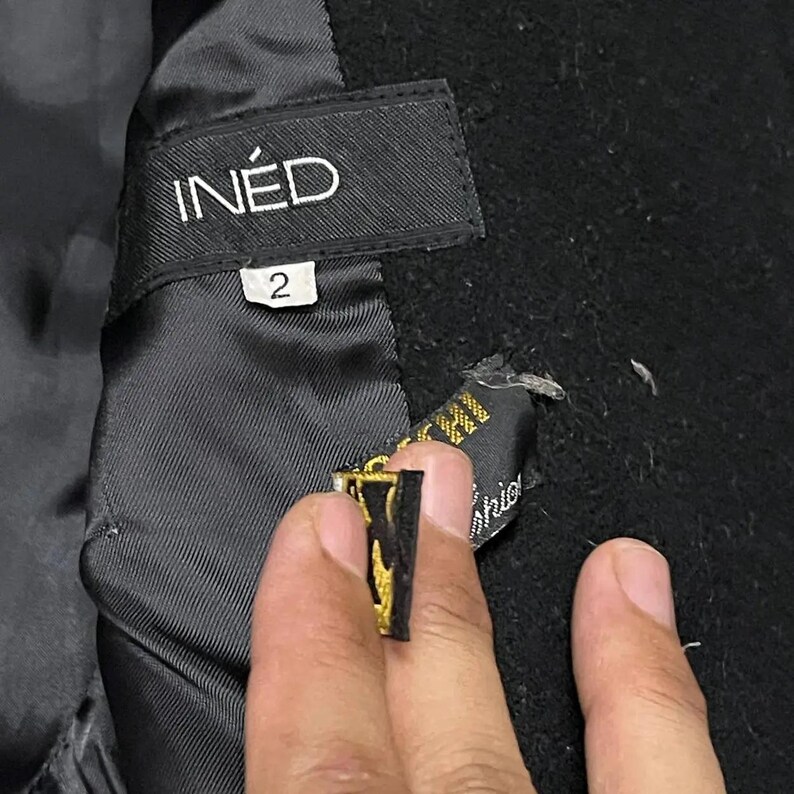 May include: Close-up of a black garment's interior, featuring a label with the brand name "IN&Eacute;D" in white lettering. Below the label is a size "2" tag. A smaller tag with gold accents is partially visible.