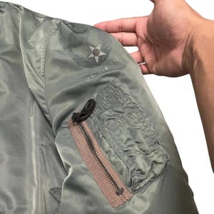 Puede incluir: Primer plano de una manga de chaqueta bomber verde oliva. Cuenta con un bolsillo con cremallera con una cremallera marr&oacute;n y una leng&uuml;eta negra. La chaqueta tiene un emblema de la Fuerza A&eacute;rea de EE. UU. en la manga superior.
